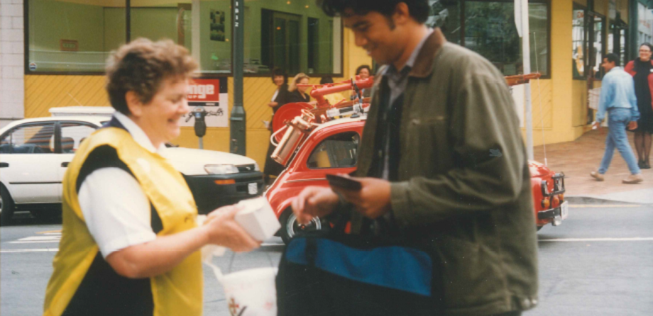 A woman in a yellow bib smiles as a man puts a donation into a bucket