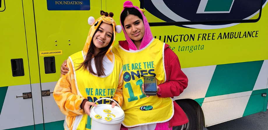 2 young women in onesies smile in front of a Wellington Free Ambulance