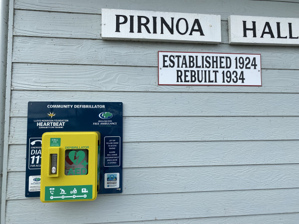 An AED installed on a light grey weatherboard building with a sign saying: Pirinoa Hall, established 1924 rebuilt 1934